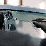 Cropped image of hands of worker in garage tinting a car window with tinted foil or film, holding special blade or knife to cut the film. Car detailing workshop, tinting windows