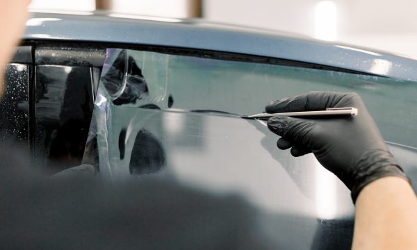 Cropped image of hands of worker in garage tinting a car window with tinted foil or film, holding special blade or knife to cut the film. Car detailing workshop, tinting windows